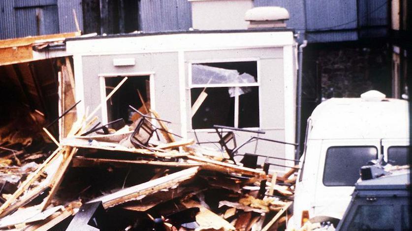 The aftermath of a bomb.  Wood and debris from the base piled up on the ground and a range rover dark blue car with an officer dressed in black standing in front of the scene. 