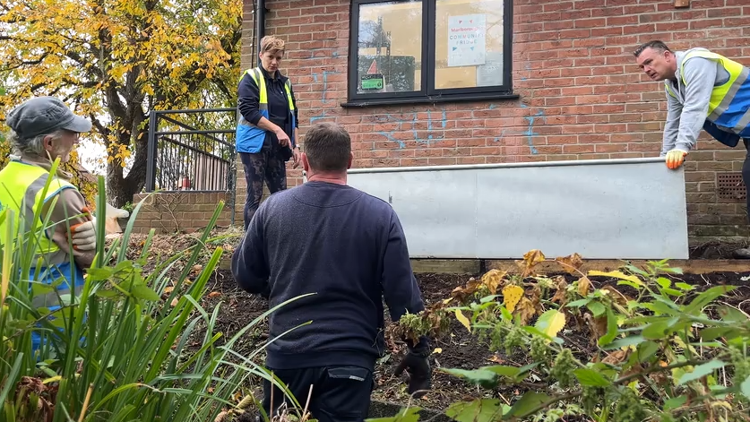 Workers and volunteers install a galvanised metal rain planter below a window, near the community fridge site and public toilets in Marlborough. Four people, three wearing yellow and blue hi-vis vests look at the positioning of the planter. 