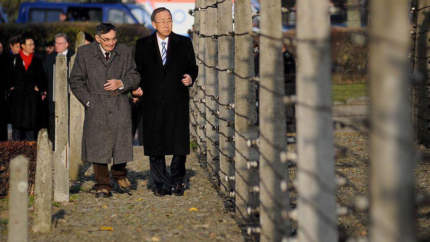 Ban Ki-moon, a tall man with short black hair and a long black coat, walks beside Marian Turski, a shorter man in a grey coat with grey hair. They are walking down a path alongside a tall, barbed fence.