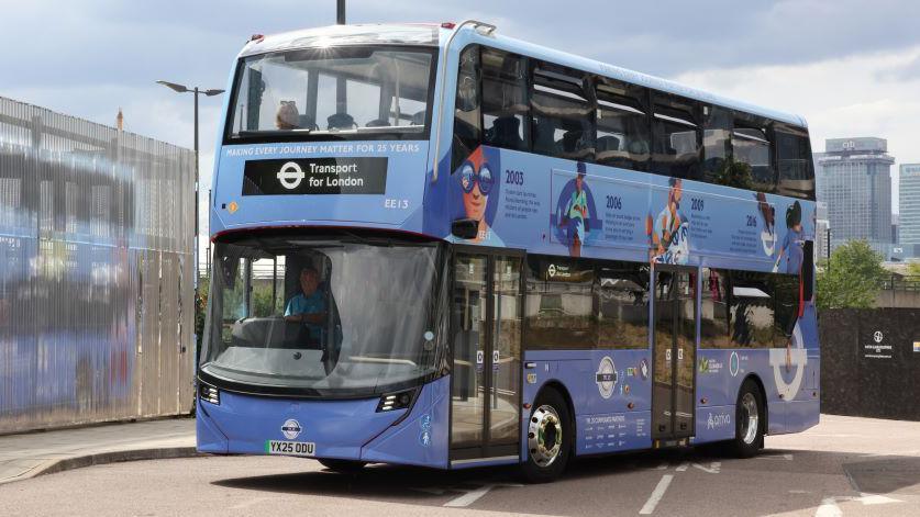A sky-blue coloured double-decker Transport for London bus is being driven along a road. It is positioned at an angle, and is viewed from the front, side-on. The bus is coloured in a gradient design, and is a darker, purple-blue at the bottom before fading to a light sky blue at the top. The bus is driven by a man in a blue shirt. The side of the bus is decorated with cartoon drawings and writing, denoting key moments in Transport for London's history. The Transport for London roundel is featured prominently across the side of the bus. The sky and some skyscrapers are visible in the distant background.