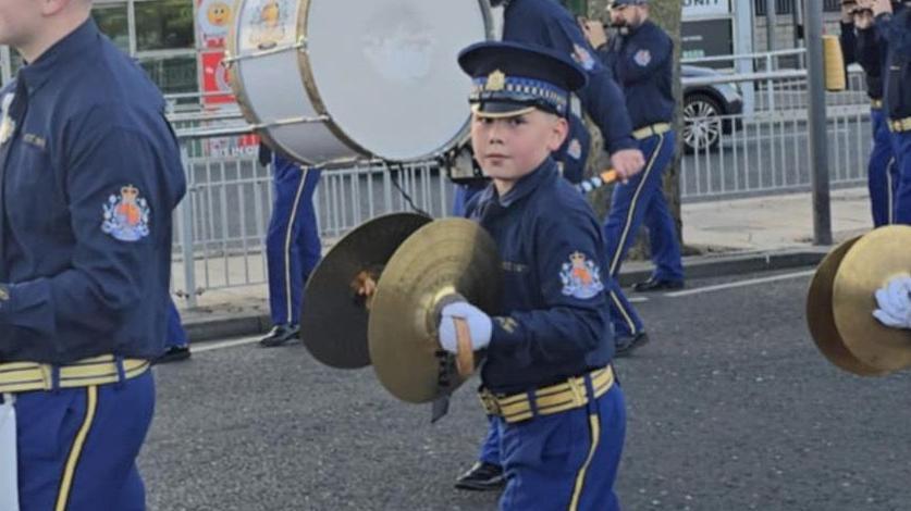 A band passes through a town. The members wear navy uniforms and are a mix of ages. Two young boys clash cymbals, while in the background a man hits a Lambeg drum
