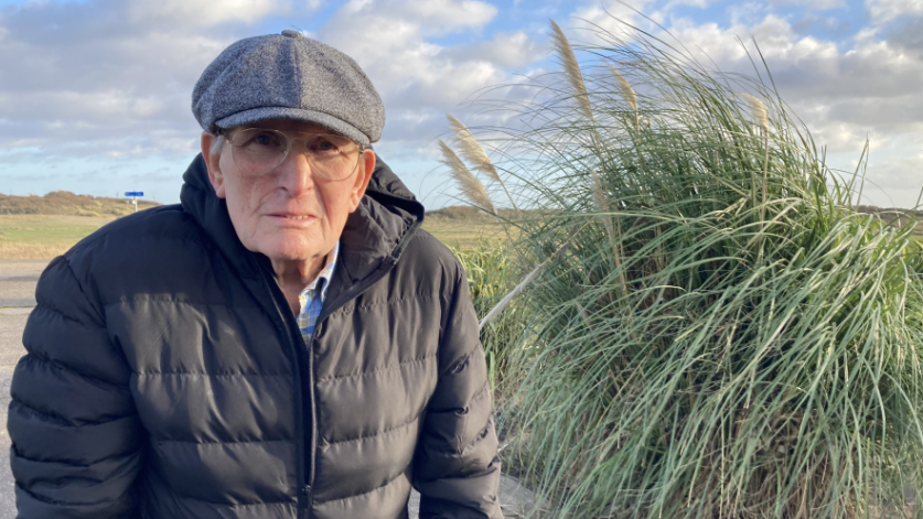 An elderly man wearing a cap and glasses with a black jacket looking directly into the camera. On the right is a green read bush. 