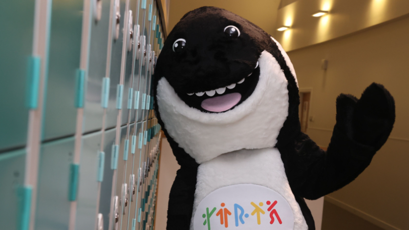 Ola the Orca standing next to a bank of lockers and is waving at the camera.