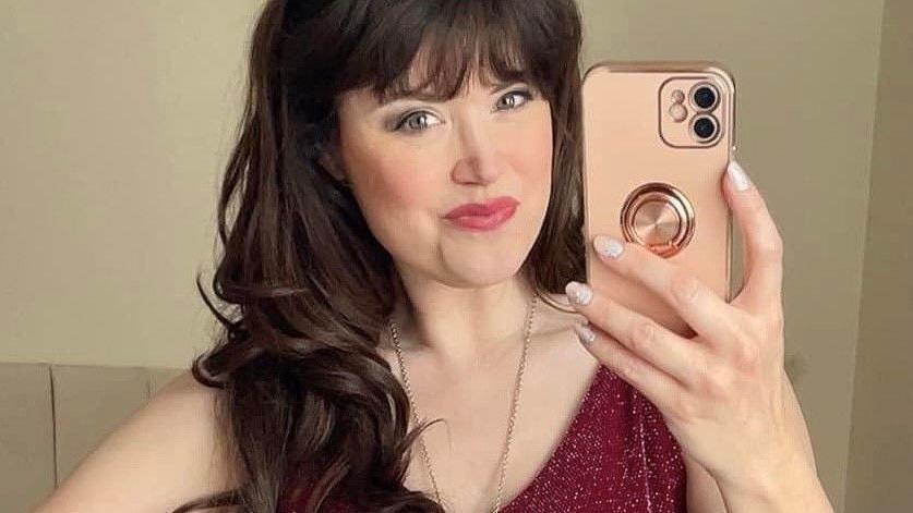 Caroline, wearing a long dark wig while holding a rose gold mobile phone in her right hand. She has a necklace on and a red sparkly top that she bought from a charity shop. 