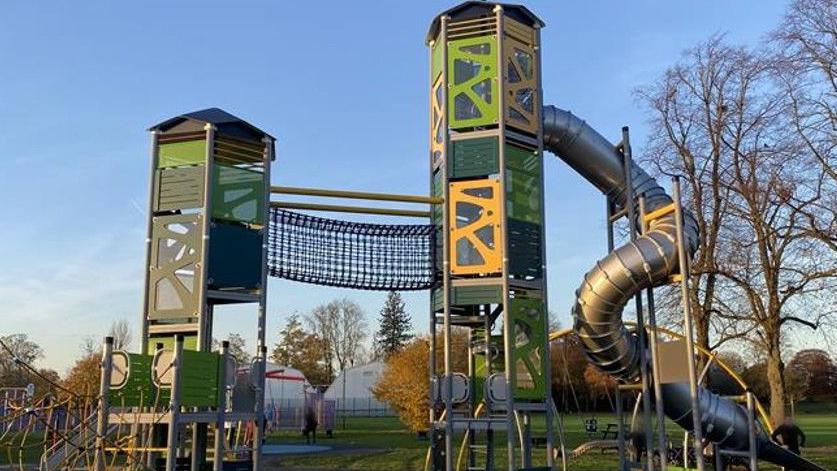 An image of a large green play area, with a large metal slide and netted bridge