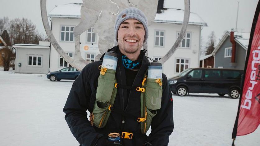 A man wearing a woolly beanie, coat, holding a can of drink and standing on snow.