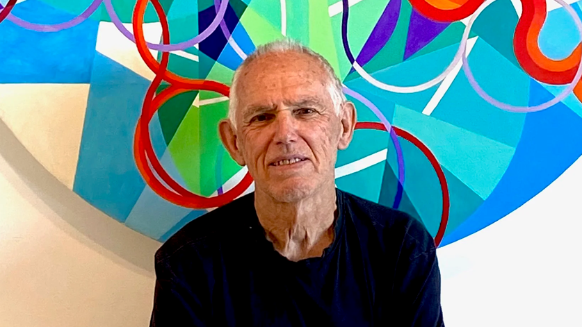 Tom Hume smiling into the camera, sitting in front of a circular geometric painting which is red, blue, white, green and purple. He has white, short hair and is wearing a dark long-sleeved top.