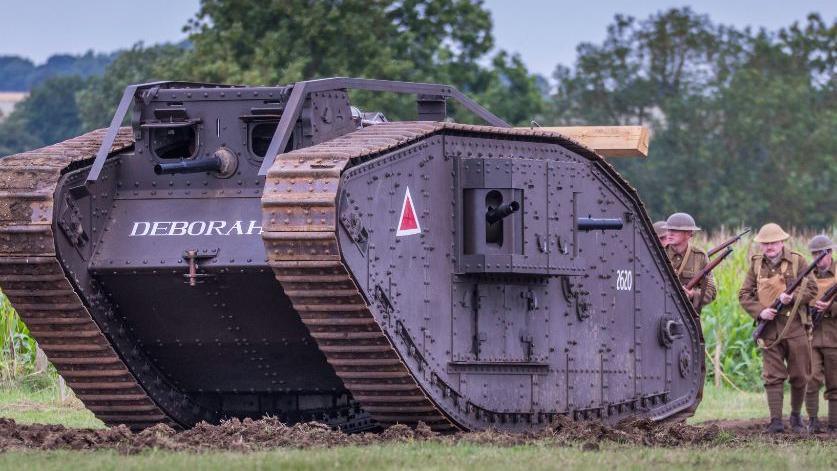 A full sized replica of a tank is pictured in a muddy field with some reenactors standing behind it wearing WW1 uniforms