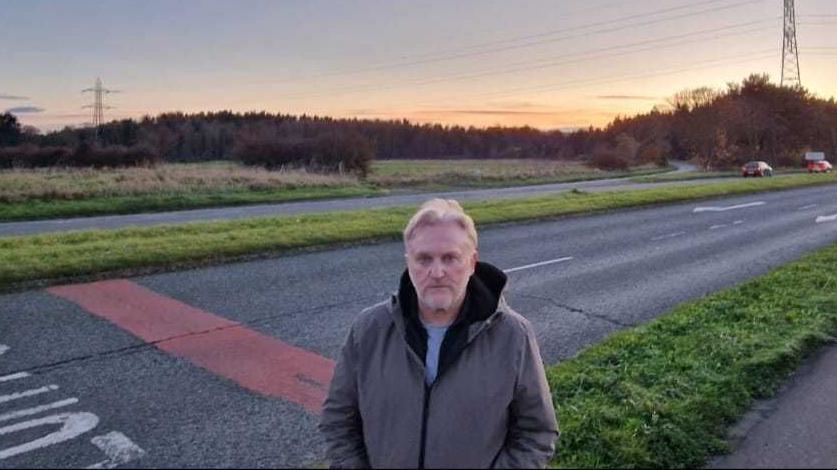 Wayne Daley looking into the camera, looking annoyed. He has light, short hair and a white beard and is wearing a green jacket with a hood. He is standing in front of a road which is surrounded by grass. Pylons can be seen on the horizon and the sun is just beginning to set.