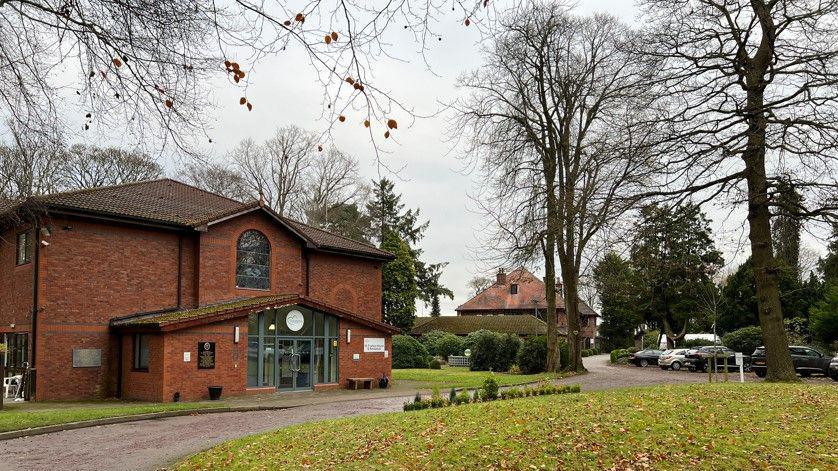 The main entrance to the hospice, with a large glass panelled entrance and a stained glass window above. Grassed areas and trees can also be seen all around, with another building and parked cars seen in the distance.