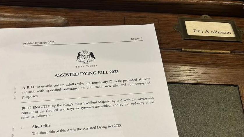 The top-half of the first A4 page of the Assisted Dying Bill 2023 has been printed and lies on the green and brown desk of Alex Allinson MHK. It bears the coat of arms of Tynwald and black writing outlines the reason for the bill.