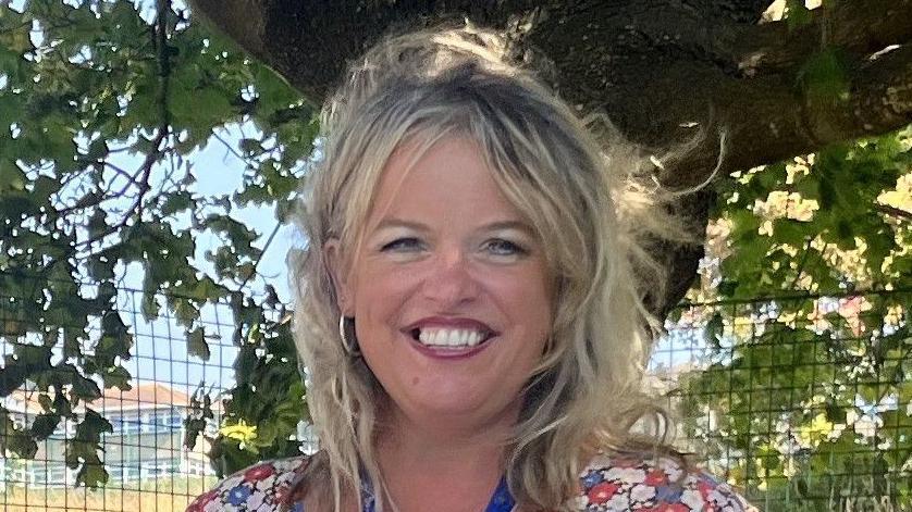 A headshot of a smiling blonde woman in a floral dress. There is a tree and a wire fence in the background