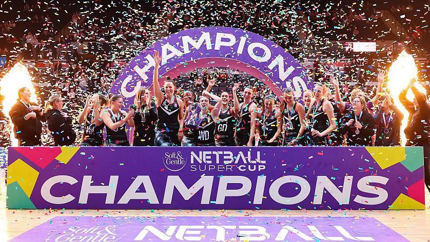Nat Panagarry of Loughborough Lightning celebrates lifting the Netball Super Cup 2026 trophy with team-mates as ticker tape flies and the team stand in front of a sign that says 'Soft & Gentle Netball Super Cup champions'