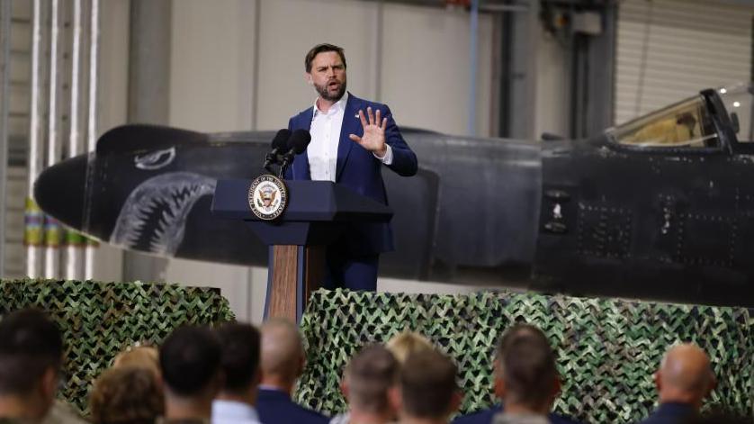 A man with dark hair and a beard makes a speech standing in front of a military aircraft with a face painted on the nose.