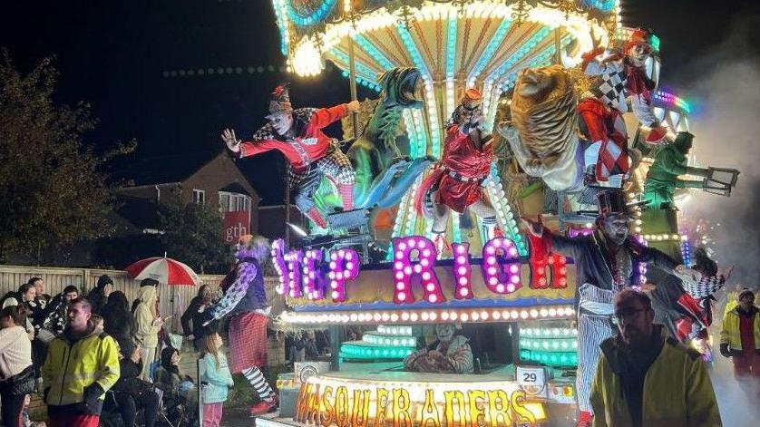 A very brightly lit carnival cart going down a street. It has dancers and figurines on it. 