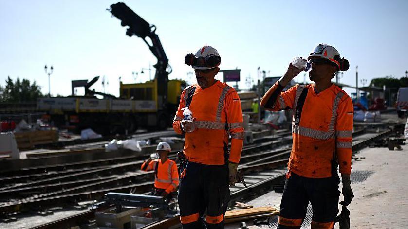 Labourers wearing hi-viz orange tops drink water to cool down as they work on a construction site of tramway lines along the Garonne river in France