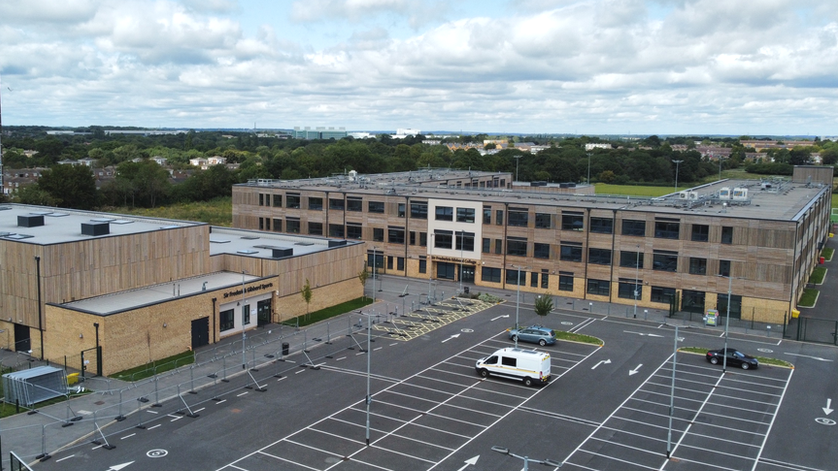 An aerial image captured of the school building and neighbouring sports hall. Both are made of light coloured brick and wood and lie next to a car park that is mostly empty. They are both fenced off. The school building is three-storey with a flat roof, while the sports hall has parts which are taller than others.