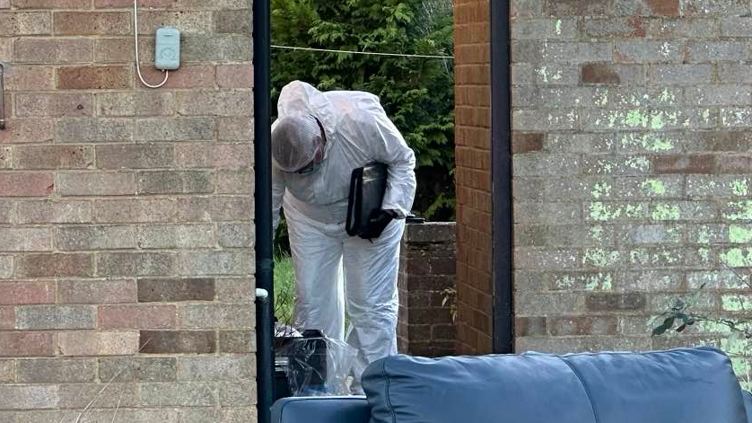 A forensic officer dressed in a white suit with a white hair net is pictured leaning over looking at something toward the ground in the side garden of a home which is behind a brick wall with an open fence door.