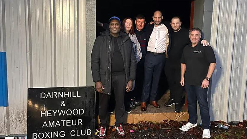 Five men and a woman who are on the committee of the community interest group raising money for the project stand in the entrance of the building with a black sign which reads: Darnhill and Heywood Amateur Boxing Club. They are all smiling.
