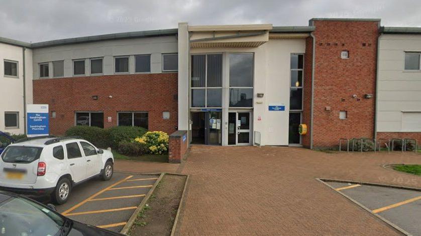 The exterior of the two-storey clinic that combines brickwork with white panelling. Large windows are visible on both levels. At the centre of the building is the main entrance, which features glass doors and a protective overhang above them.
To the left of the entrance, a sign identifies the building as The Sandringham Road Clinic. In front of the building, several vehicles are parked in designated spaces, including a white SUV. The surrounding area is tidy and landscaped, with bushes and greenery near the entrance. The sky is overcast.