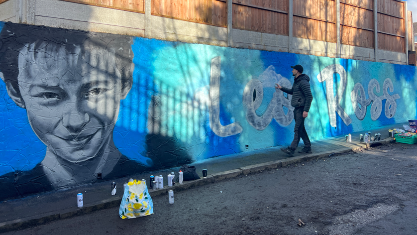 A man touches a wall on which a gigantic blue mural has been painted. On the left is Leo Ross's face, neck, and shoulders. On the right is his name, painted on the wall in white writing.