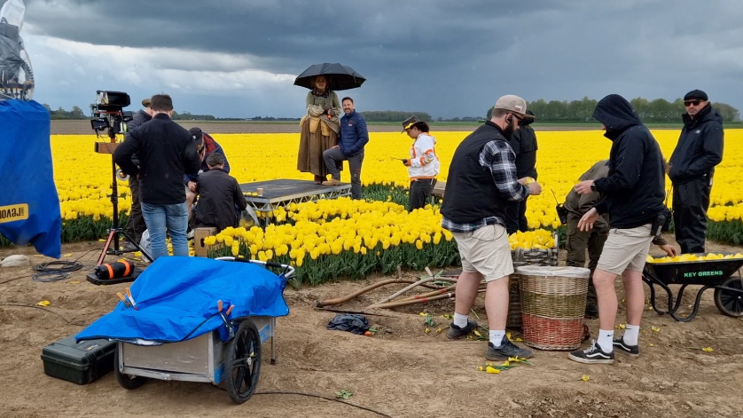 Film crew in a field of yellow tulips, surrounded by various filming equipment including a camera and small trailer. A woman in period costume is in the centre of the image, holding an umbrella