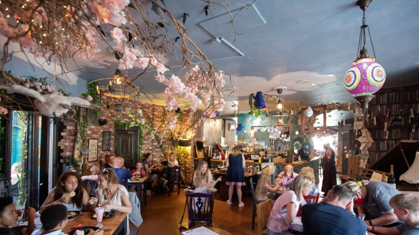 There are several adults and children sitting at tables in a cafe, which is decorated with branches of blossom, trees, fairy lights and colourful lamps
