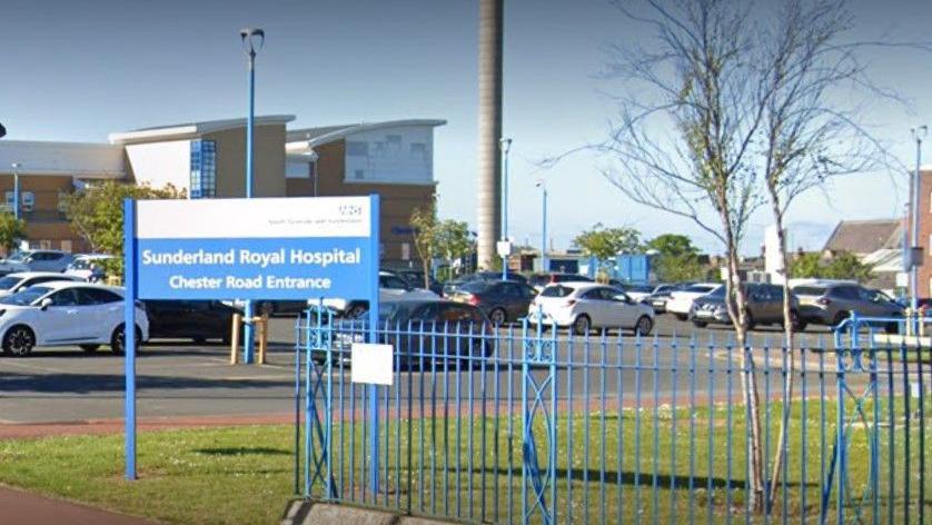A blue sign reading Sunderland Royal Hospital Chester Road Entrance. There is a car park behind it, with the hospital's tall beige and grey buildings further back. A blue fence is separating the grassland near the car park from the street pavement. 