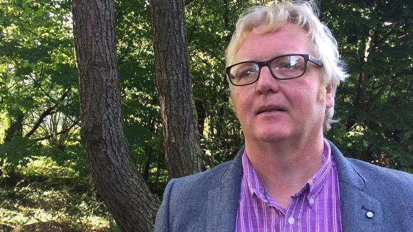 James Orr - a man with white hair and black rimmed glasses stands in front of a tree. He is wearing a blue blazer and a purple striped shirt.