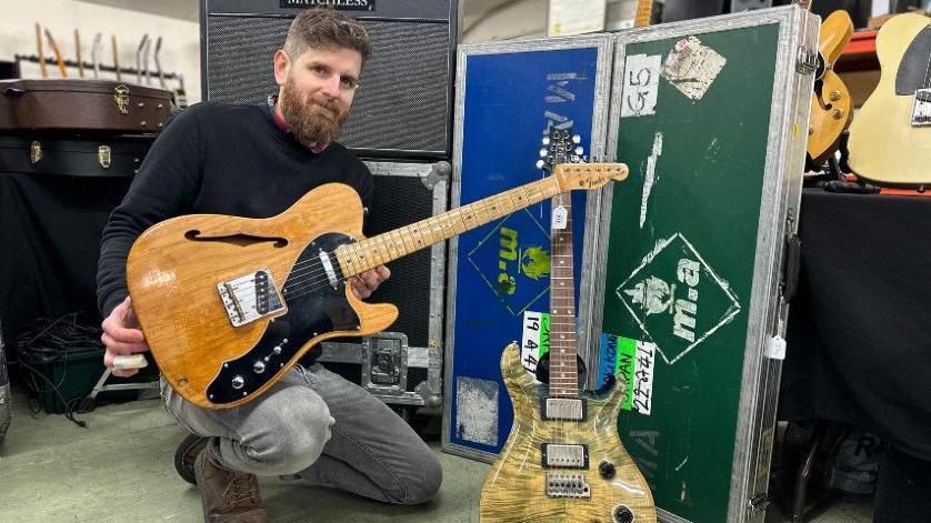 Auctioneer Luke Hobbs on one knee holding one of the guitars which is on sale. He is wearing grey jeans and a black jumper. Another of the guitars is on a stand.