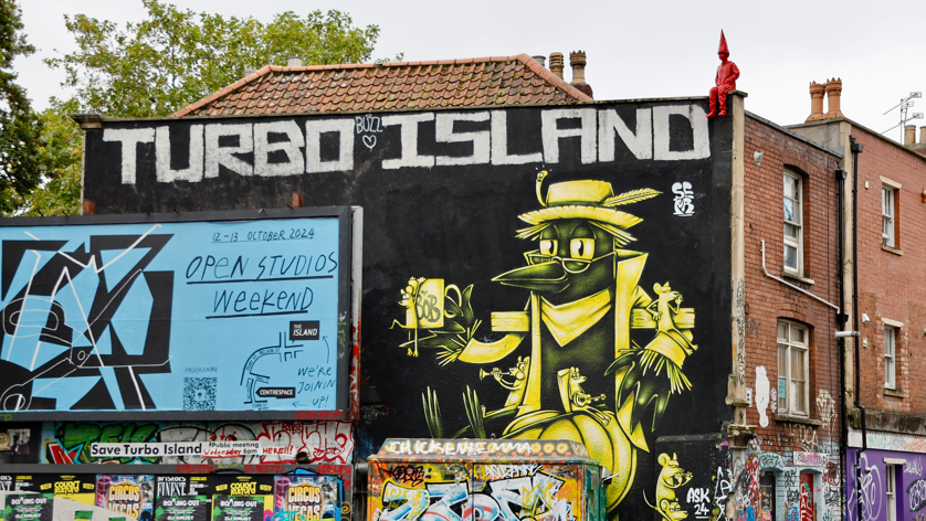 A red sculpture of a boy wearing a long pointed 'dunce' hat can be seen perched on the edge of a city building. The building has a mural of a yellow bird on the side and is painted black, with the words "turbo island" in large block letters above it. 