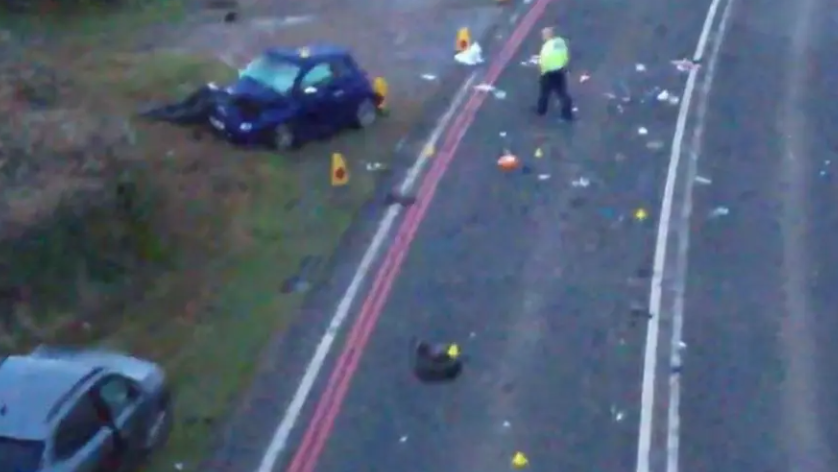 The scene of the crash with two cars off the road on the verge, one with a severely damaged bonnet. There are police cones around this car and a police officer in a high-vis jacket walking towards it. Other debris is scattered on the tarmac.