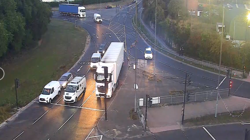 Several vehicles, including a white van and a white lorry, appear to wait at traffic lights at a roundabout. It is dimly lit because it is early morning.