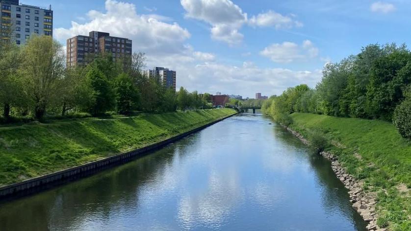 A river is surrounded by green grass and trees as well as apartment buildings. 