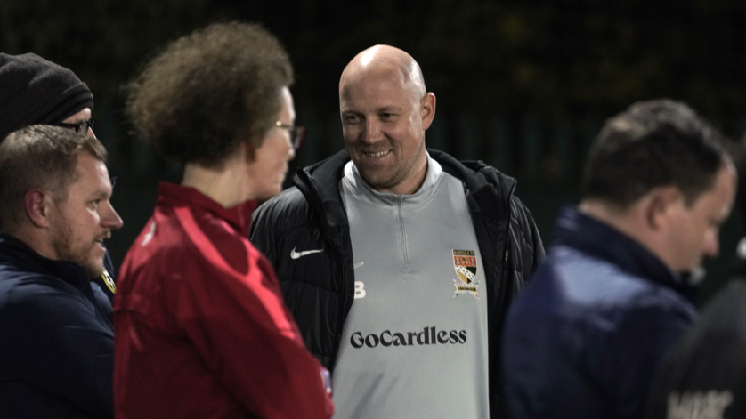 James Buhlemann is wearing a black coat over a grey Norfolk FA top. He appears to be standing next to a pitch with some other men.