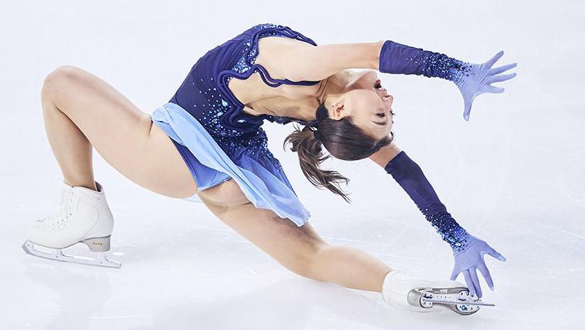 Japan's Kaori Sakamoto a figure skater in a blue costume performs a deep lunge on ice, arching backward with one arm extended and the other reaching down, against a smooth white background.