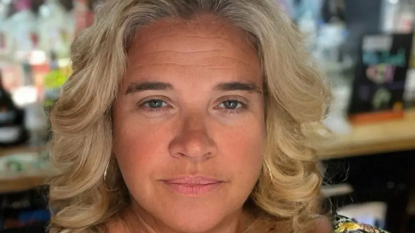 Miranda Richardson smiles at the camera. She is standing behind a bar with bottles of alcohol behind her. She has curly blonde hair and is wearing hoop earrings.