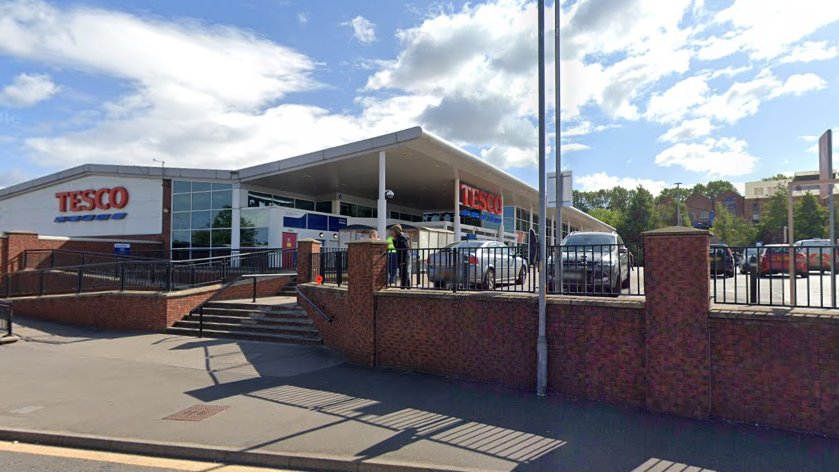 A view of Tesco superstore from the road. The store has a white roof and glass front and has Tesco in red capital letters on both the front and side. There is a brick wall around the side with black railings.