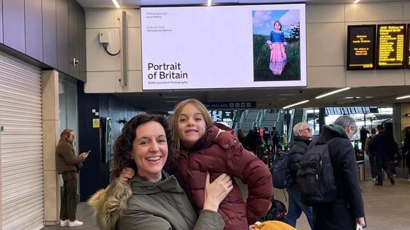 A dark-haired smiling woman holds up her six-year-old daughter in Leeds Station. They stand in front of a board, which displays a picture of the girl in an Indian dress with the words "Portrait of Britain" next to it.