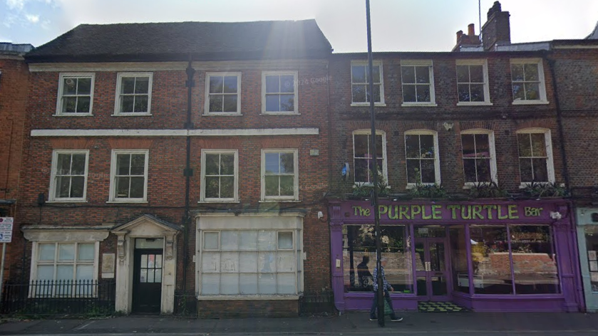 Google Street View image of the 18th Century office building and the Purple Turtle from across Gun Street. They are both three storeys tall and the Purple Turtle is painted purple. 