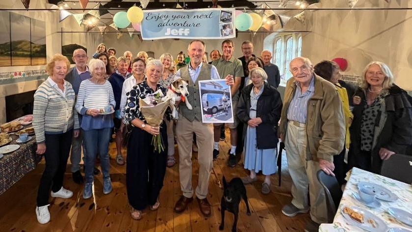 Somerset newspaper delivery man Jeff Savage stands inside a large hall, surrounded by friends, holding a dog and a photo of himself at work. A banner in the background reads "enjoy your next adventure Jeff", while a woman next to him holds a bouquet of flowers. On the far left of the photo sits a table covered with cake and other sweet treats.