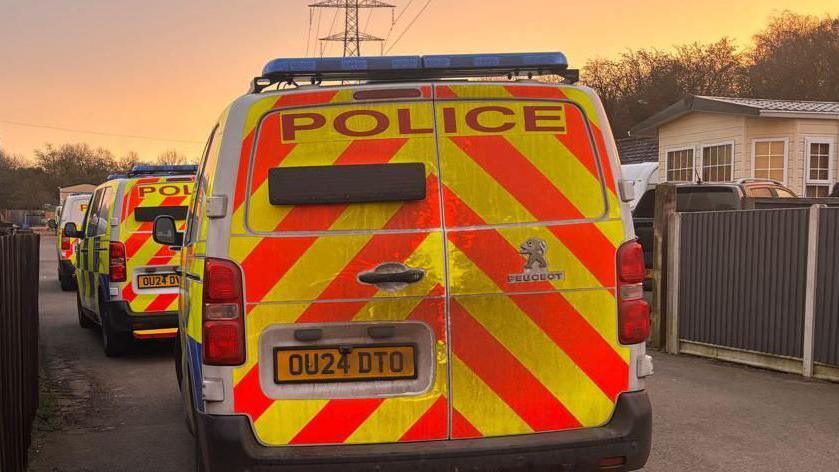 A rear view of three police vans lined up, one behind the other, on a concrete driveway, with a mobile home on the right.