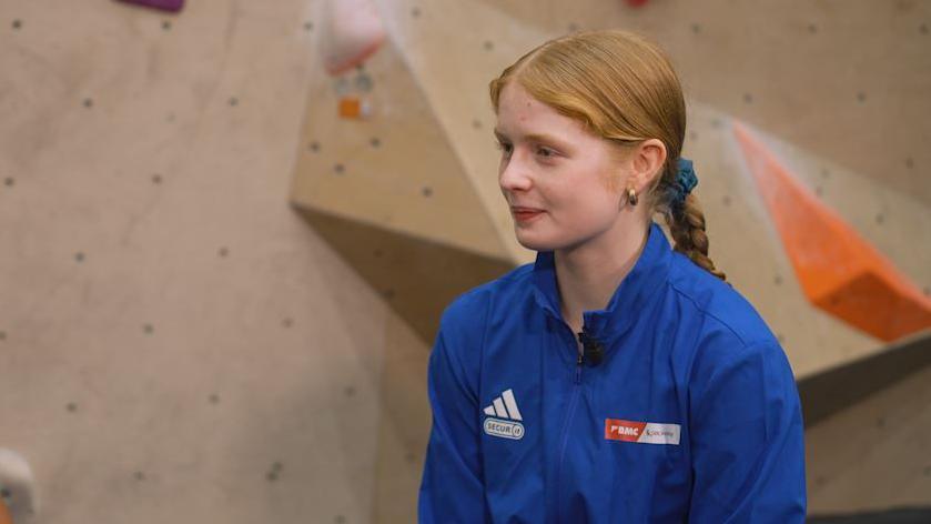 Climber Connie Bridgens sits in front of a colourful climbing wall, wearing a blue jacket.