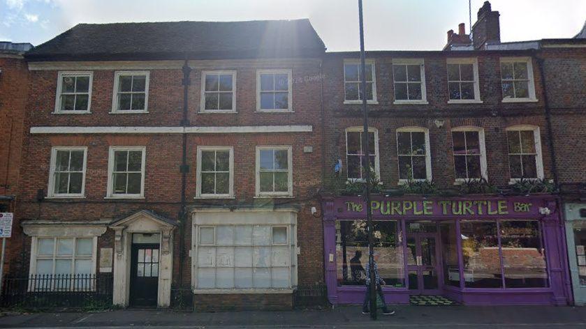 Google Street View image of the 18th Century office building and the Purple Turtle from across Gun Street. They are both three storeys tall and the Purple Turtle is painted purple.
