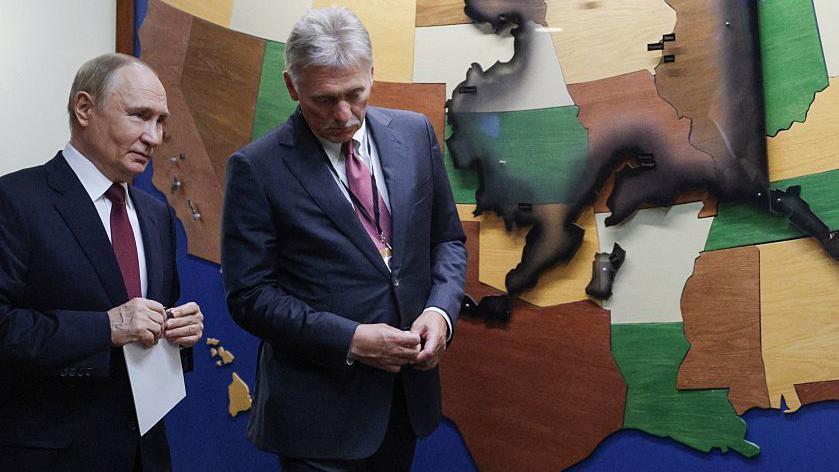 Two men in suits stand with a backdrop showing American states including Alaska.