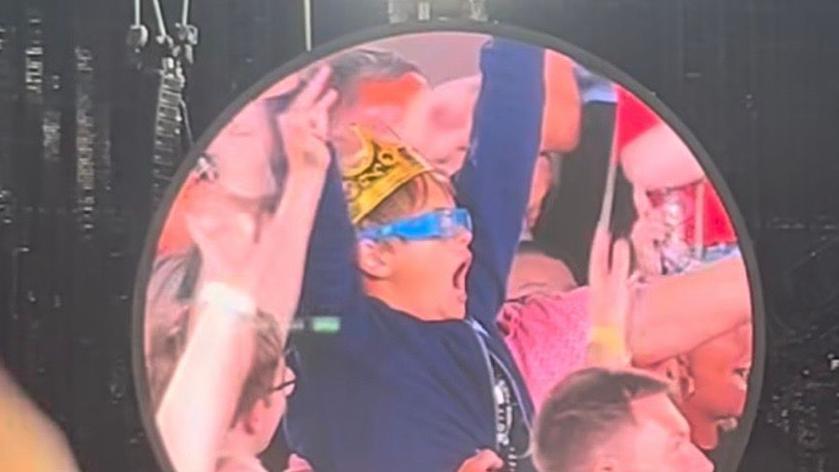 A circular screen is fixed to a black stage. In the middle of the screen is Issac wearing a gold crown and a blue hoodie and glasses. He has both hands in the air and people are sitting around him. He appears to be cheering.