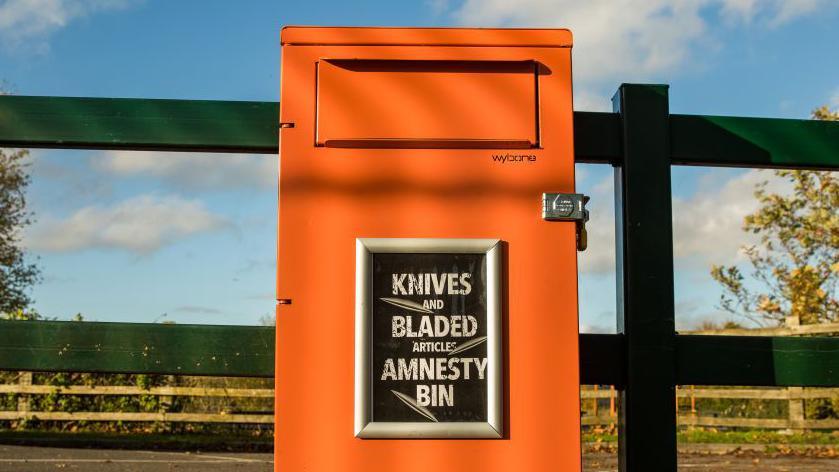 An orange metal bin against a green wooden fence with the word's knives and bladed articles amnesty bin written in white against a black background attached to the front of the bin