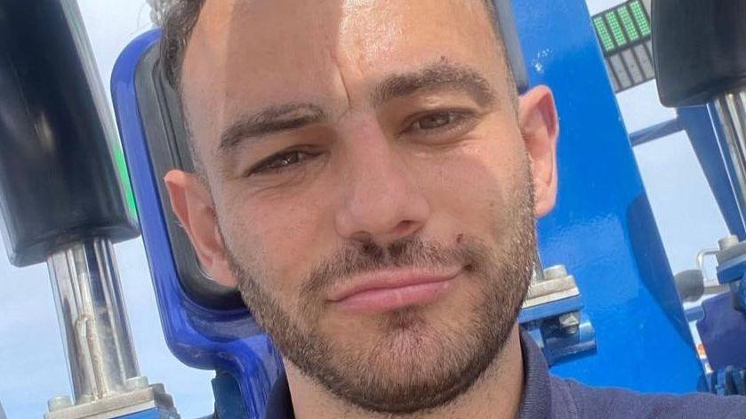 Corrie Lee Stavers takes a selfie. He is a young man with short dark hair and neat thin dark beard. He is wearing a navy polo shirt and appears to be in the seat of a blue funfair ride.