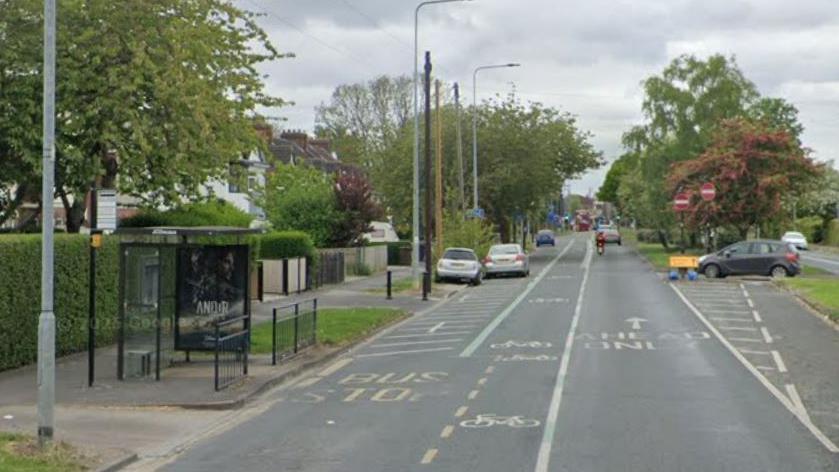 A bus shelter next to a tree-lined road with residential housing on the left of the picture