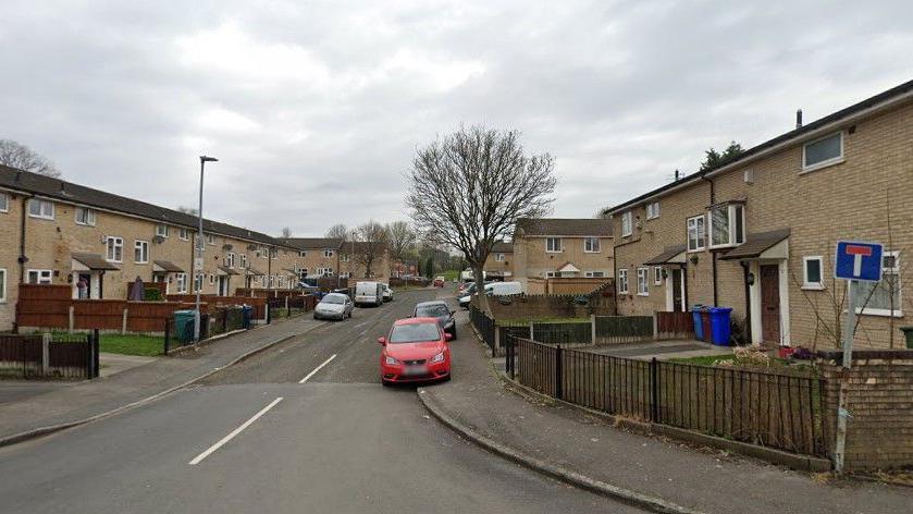 Bob Massey Close. There are terraced brick-built houses either side of the road. A number of cars are parked and several trees can be seen.